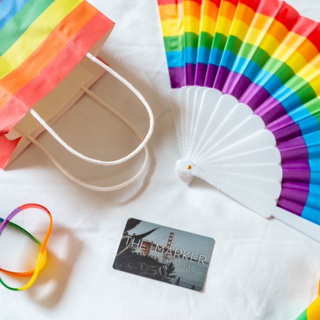 marker hotel key card on a bed with gay pride goodies: fan, lei, wristbands, and a bandana